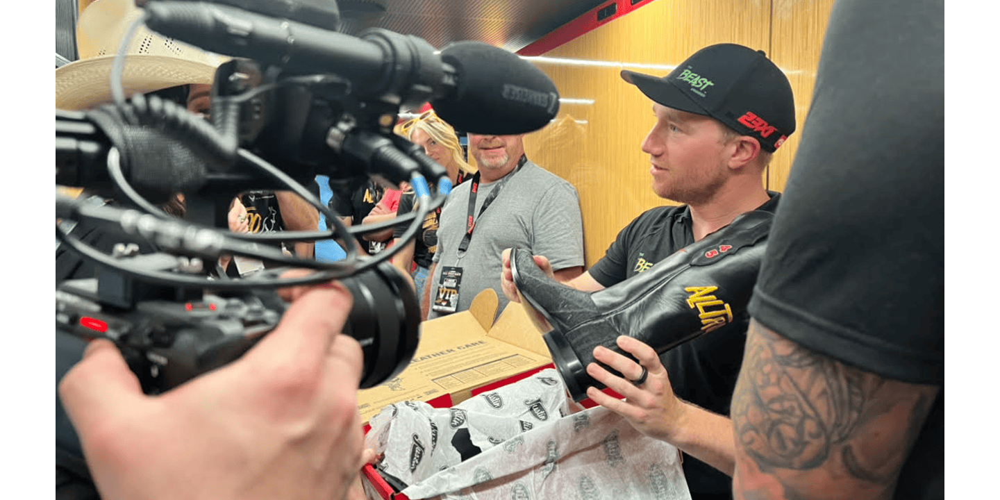 El piloto de 23XI Racing, Tyler Reddick, con una gorra y una camiseta de béisbol negras, muestra sus nuevas botas Justin personalizadas ante una cámara en la NASCAR All-Star Race mientras también sostiene una caja de botas Justin roja.